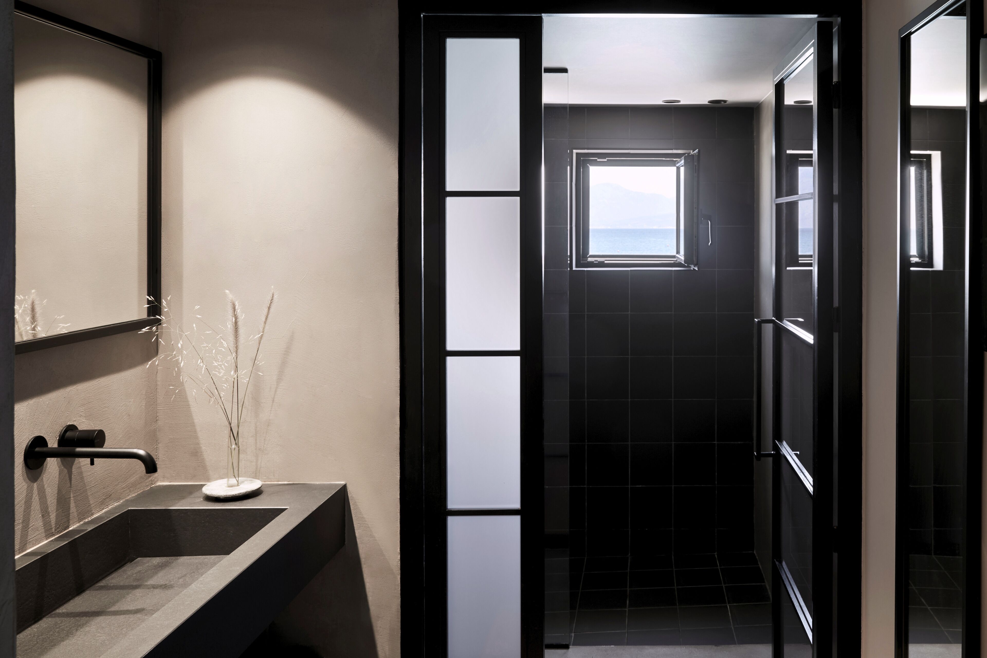 Un baño moderno con un espejo grande, un lavabo oscuro y vista al mar a través de una ventana.