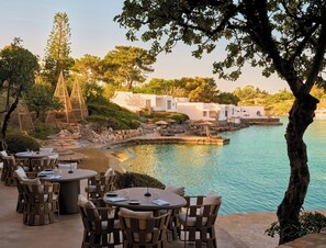 An outdoor dining area with round tables and chairs, overlooking a body of water and a landscaped garden.