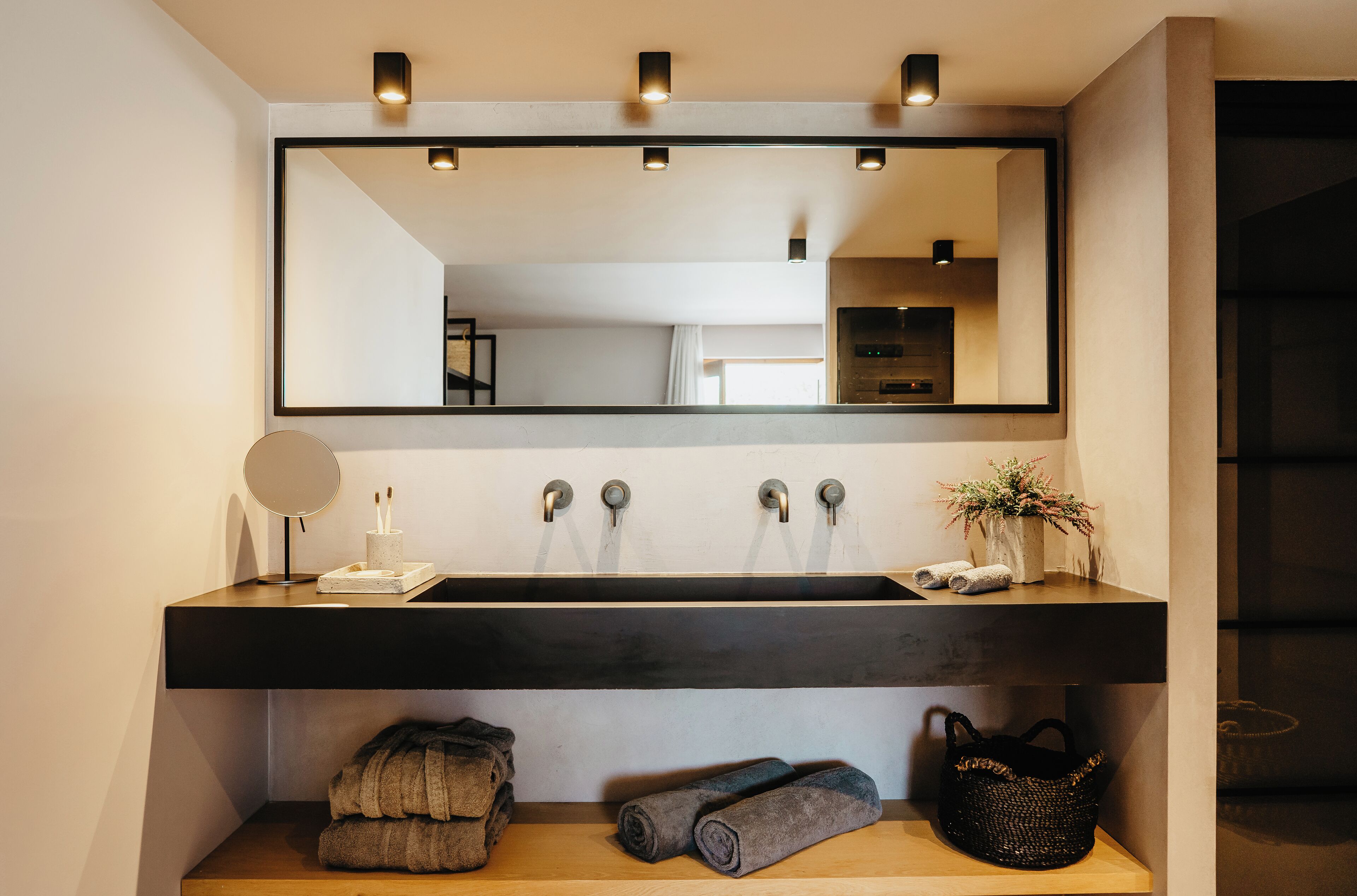 A modern bathroom with a large mirror, a dark countertop, and a wooden shelf with towels and a basket.