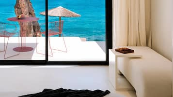 A hotel room with a bed, a small table with a fruit bowl, a red chair, a thatched umbrella, and a view of the ocean.
