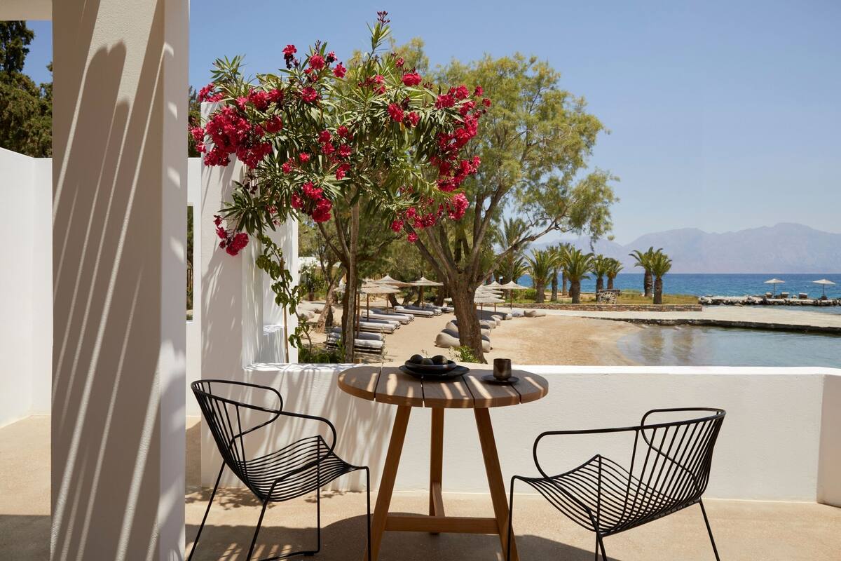 Una terraza con dos sillas de alambre negras, una mesa redonda de madera y vistas a la playa y a las montañas.