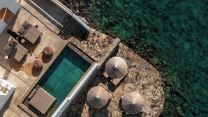 Vista aérea de un área de piscina con sillones reclinables, sombrillas y una terraza de piedra con vistas a aguas cristalinas.
