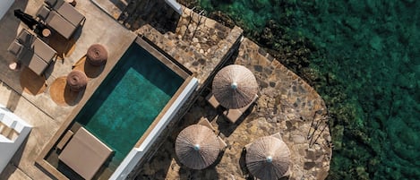 An aerial view of a pool area with lounge chairs, umbrellas, and a stone deck overlooking clear water.