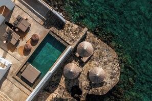 An aerial view of a pool area with lounge chairs, umbrellas, and a stone deck overlooking clear water.