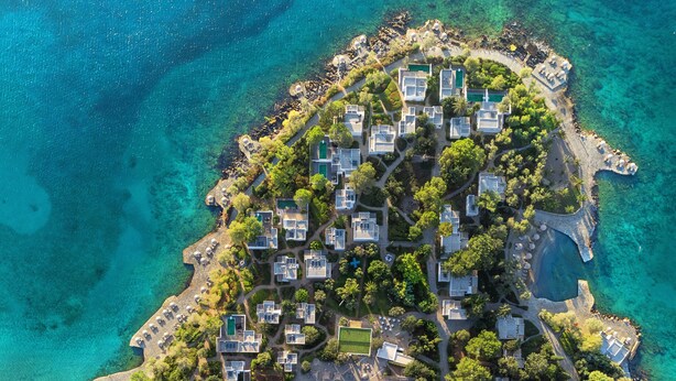 Vista aérea de un complejo turístico costero con varias villas, una piscina central y aguas turquesas cristalinas.