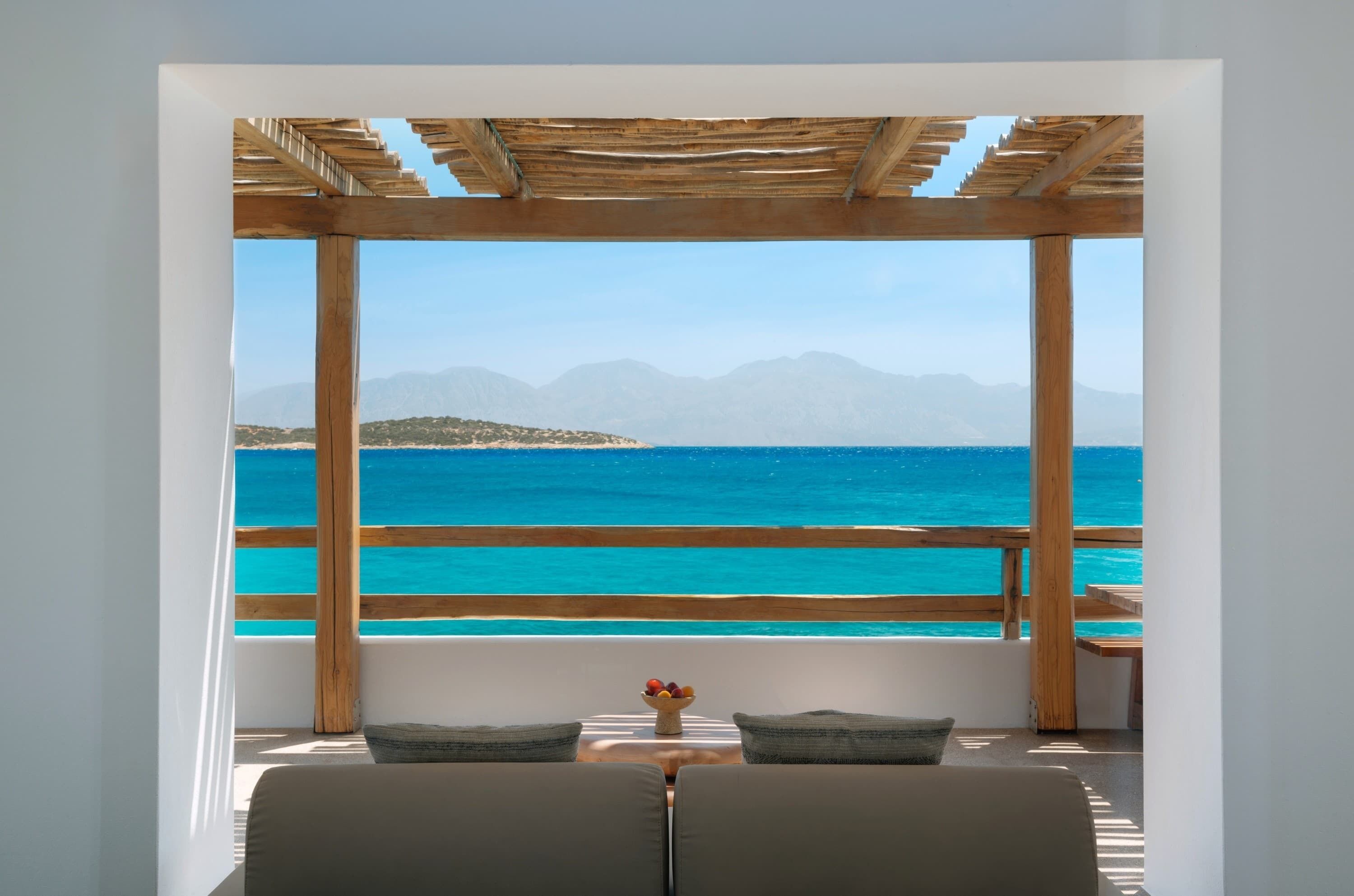 A room with a view of the sea and mountains, featuring a wooden pergola, a sofa, and a small table with fruit.