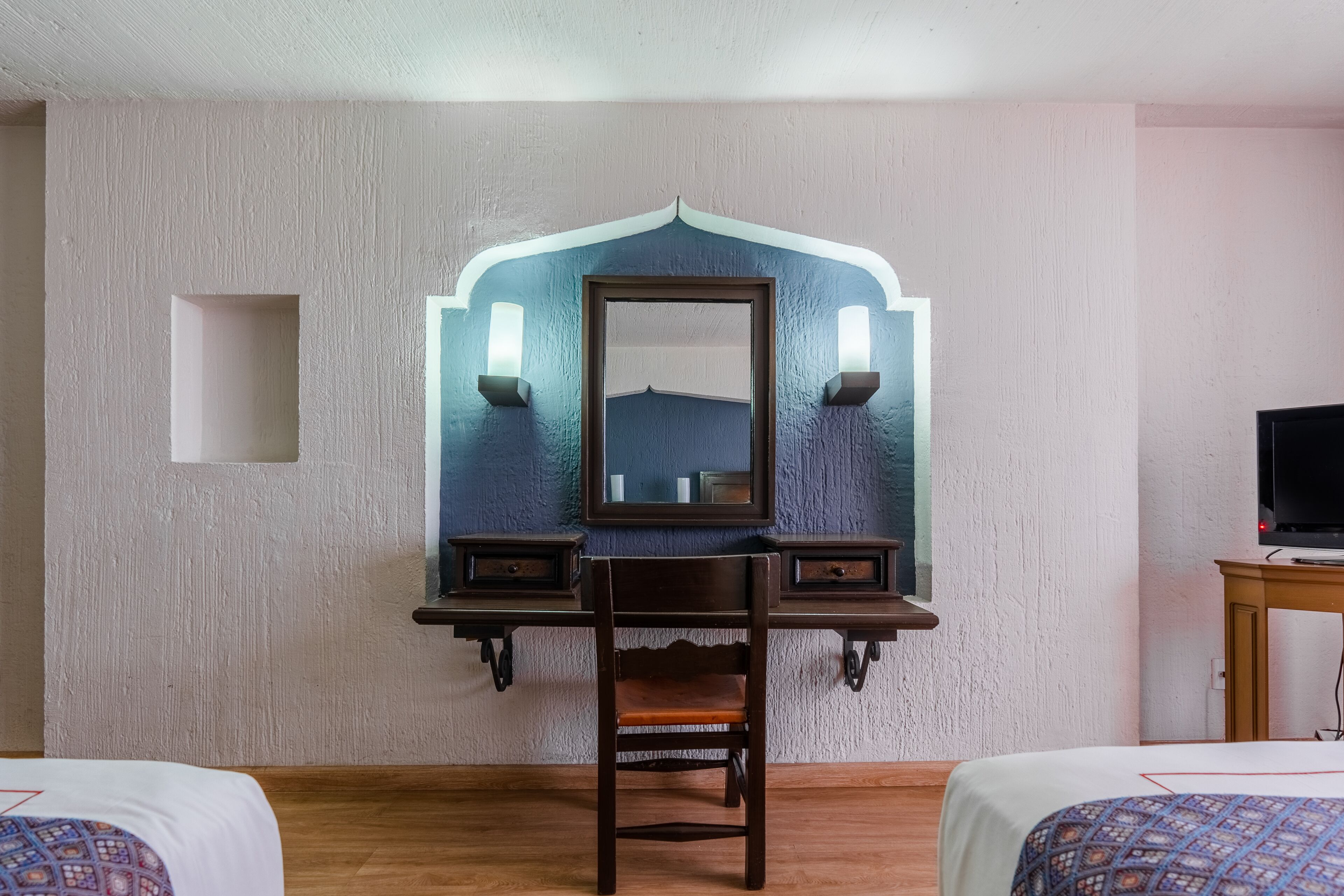 A hotel room with a wooden desk, a chair, a bed with patterned bedding, a television, and a mirror with wall sconces.