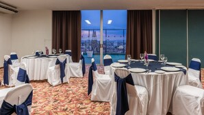A conference room with round tables, white chairs with blue sashes, and a view of a cityscape through large windows. - Hotel Fenix (Guadalajara)