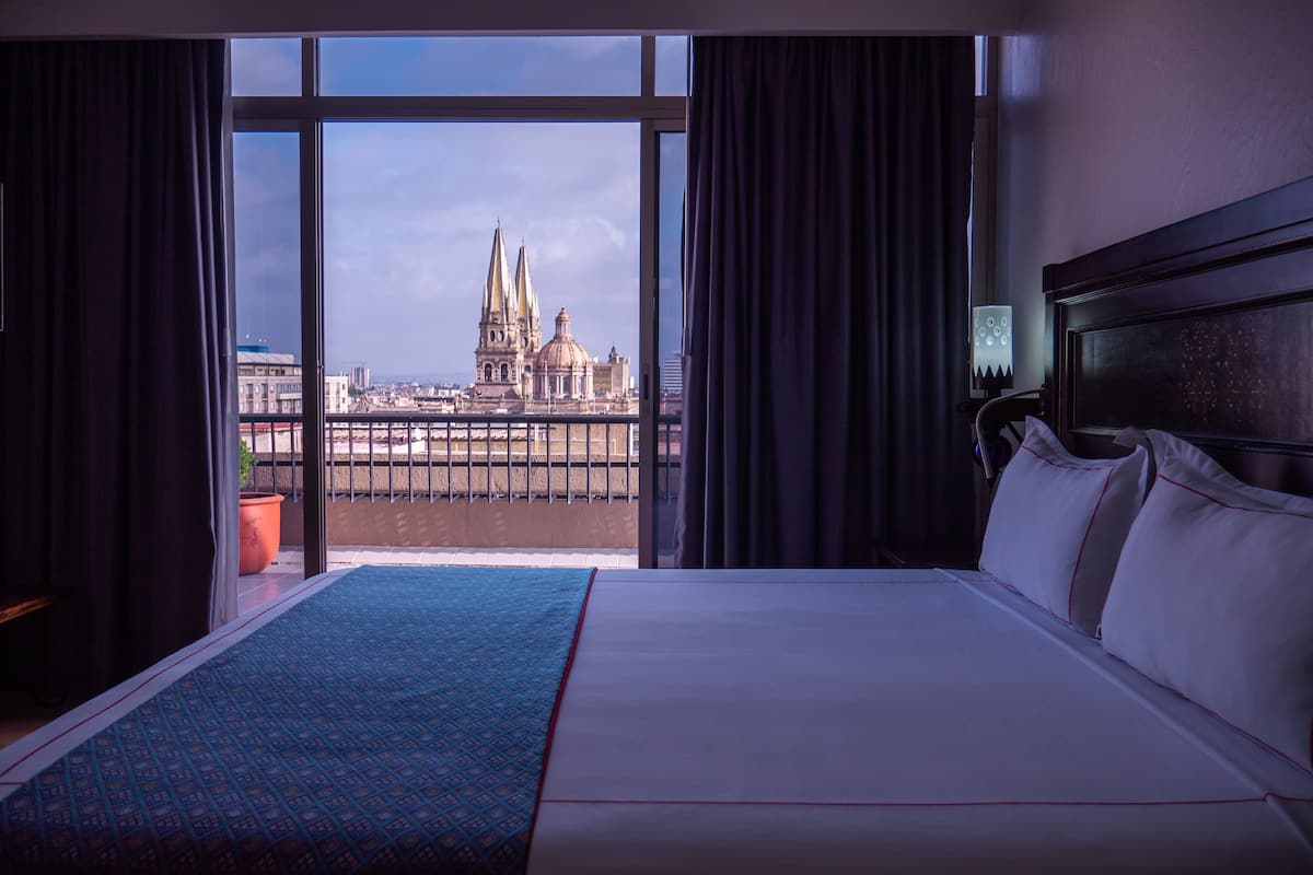Habitación de hotel con una cama grande, vistas de una catedral a través de una ventana y un balcón con barandilla.