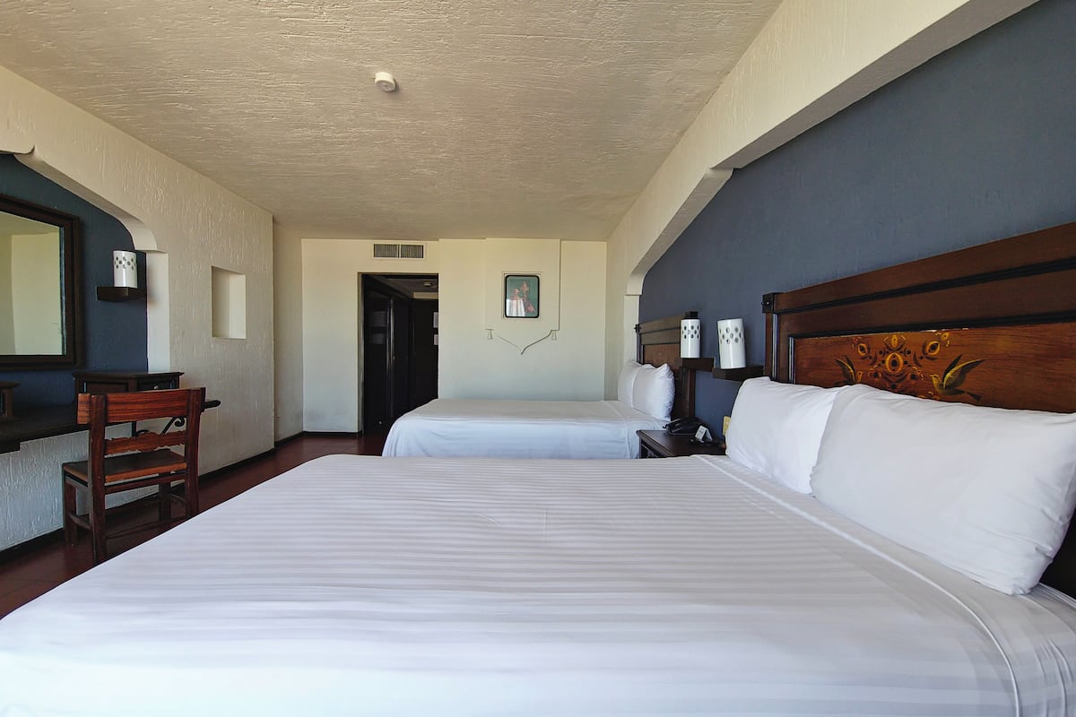 Habitación de hotel con dos camas, cabecera de madera, escritorio, silla, espejo y una puerta.