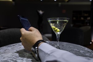 A hand holding a smartphone with a martini glass containing olives on a marble table.