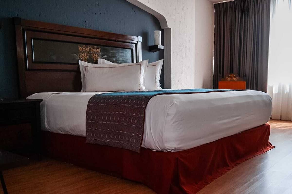 Habitación de hotel con cabecera de madera, cama grande con falda roja, mesita de noche y mesilla auxiliar.