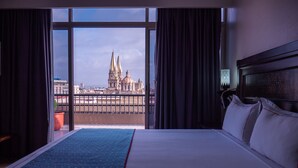 A hotel room with a large bed, a view of a cathedral through a window, and a balcony with a railing. - Hotel Fenix (Guadalajara)