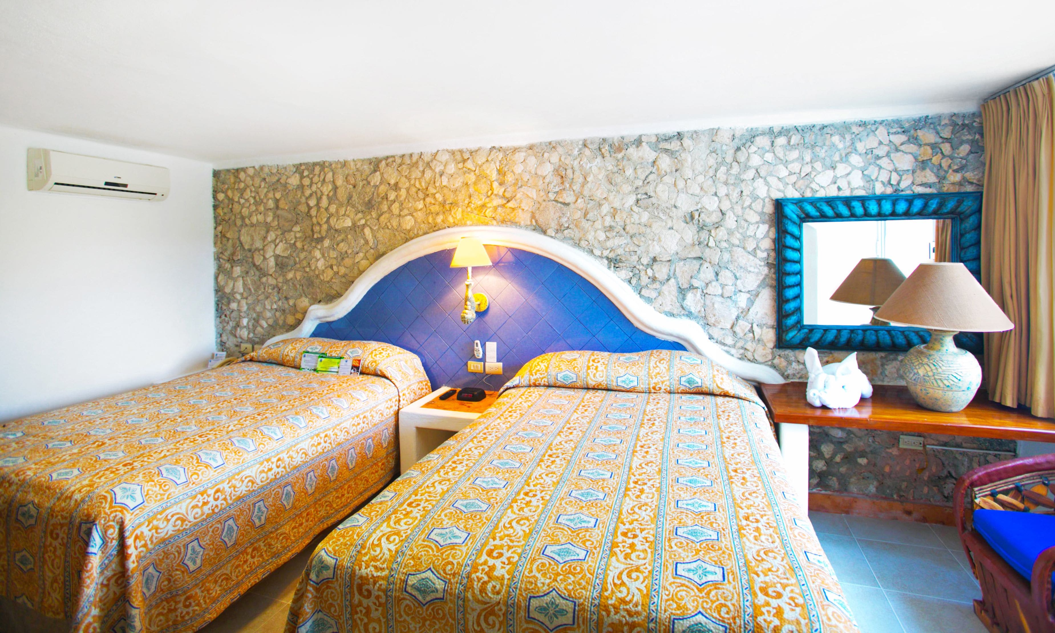 Ein Hotelzimmer mit zwei Betten, einer Steinsäule, einem Spiegel, einer Lampe und einem kleinen Tisch mit Fernbedienung.