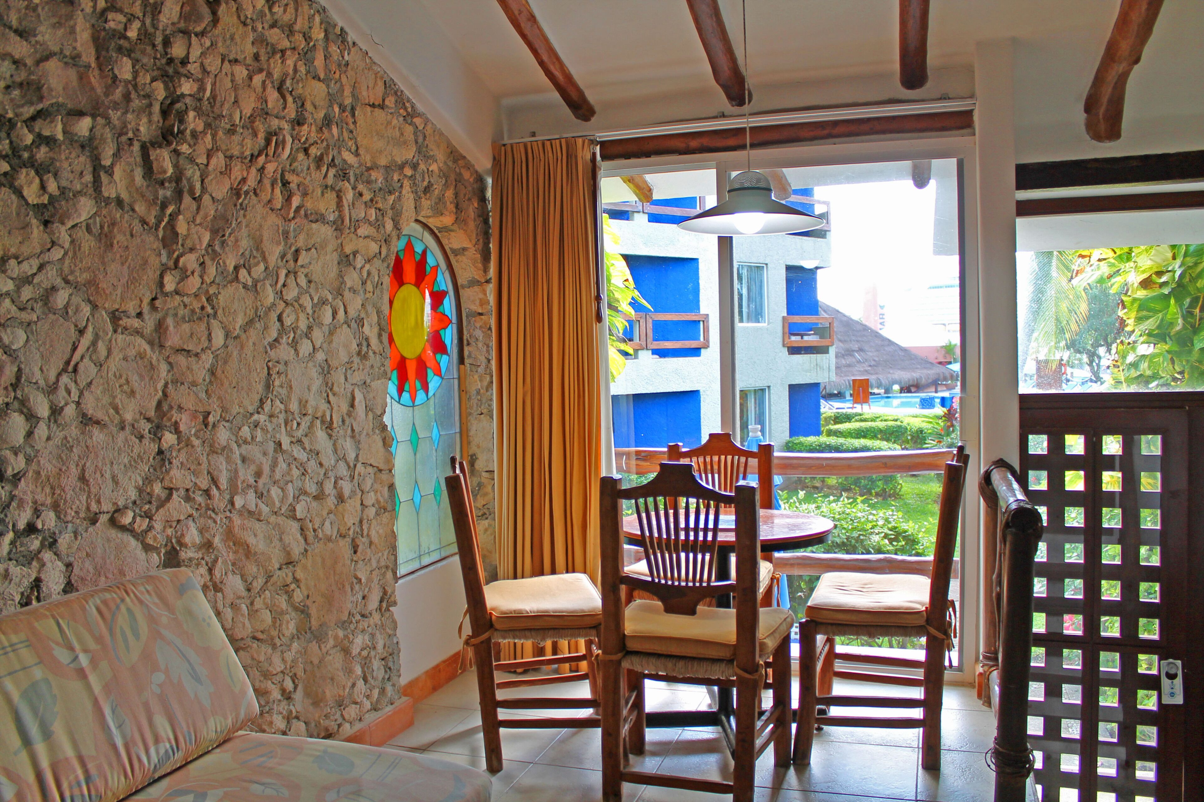 A room with a stone wall, a wooden chair, a cushioned bench, and a view of a building with blue balconies.