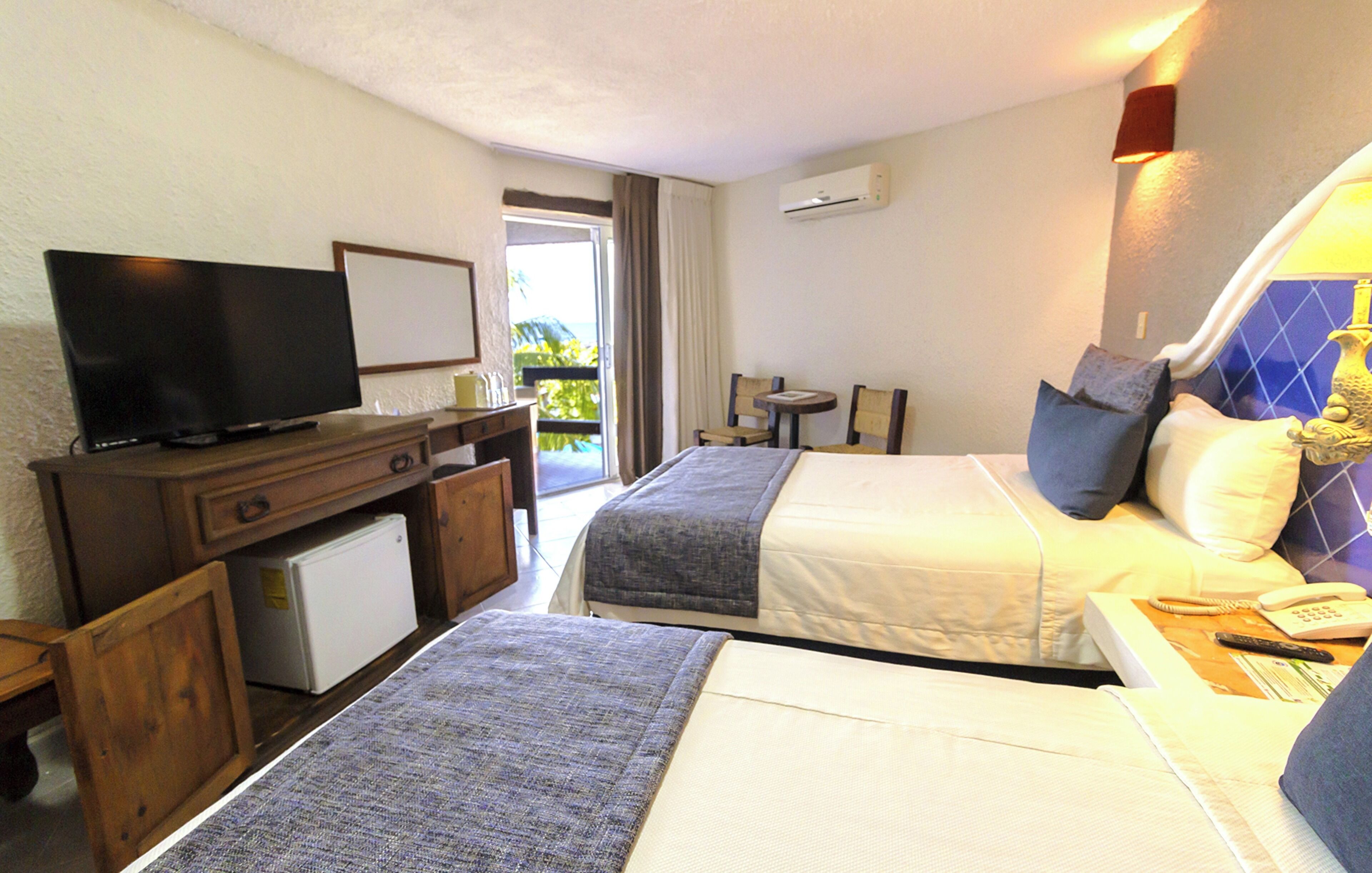 Quarto de hotel com duas camas, uma escrivaninha com televisão, um pequeno frigorífico e uma varanda com vista.