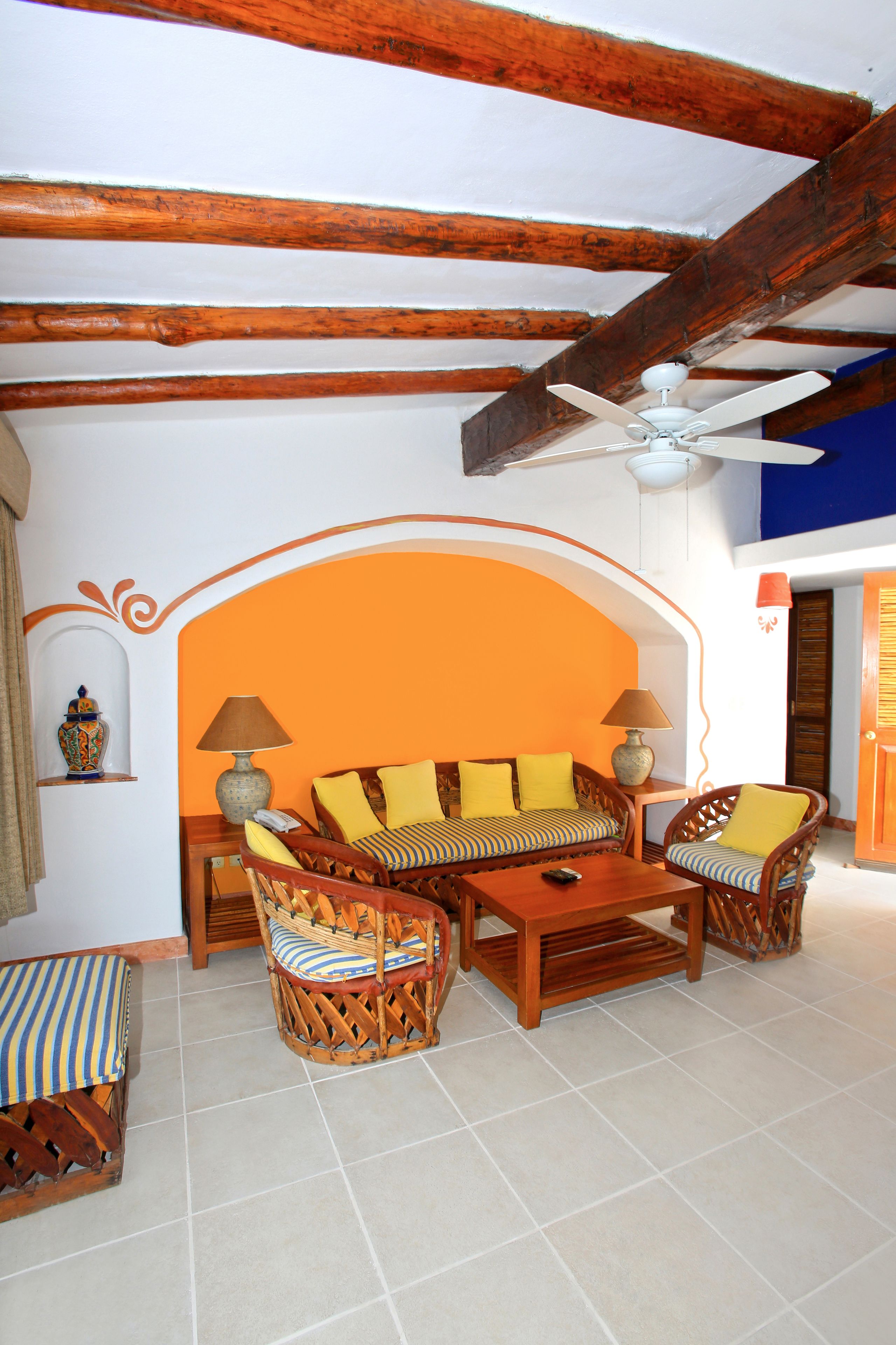 A room with a wooden ceiling, a sofa set with yellow cushions, a wooden coffee table, and a small table with a lamp.