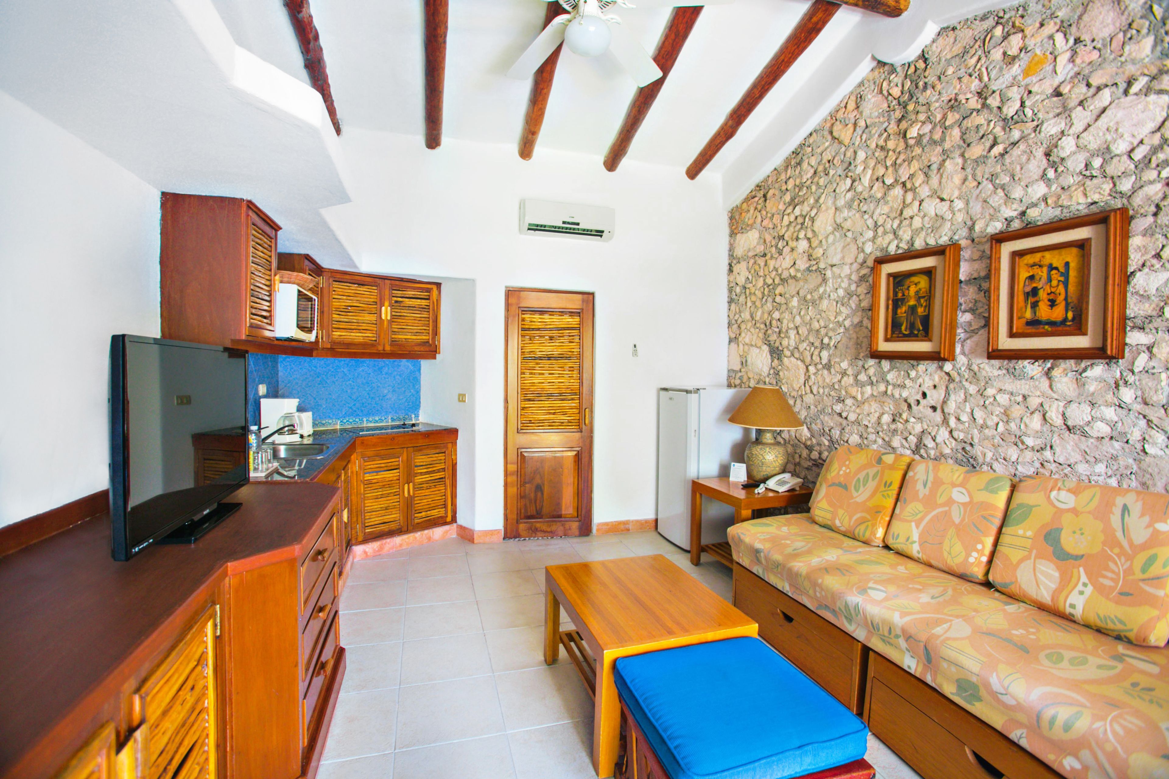 A compact living space with a kitchenette, a sofa with patterned cushions, a wooden bench, and a stone wall with framed artwork.
