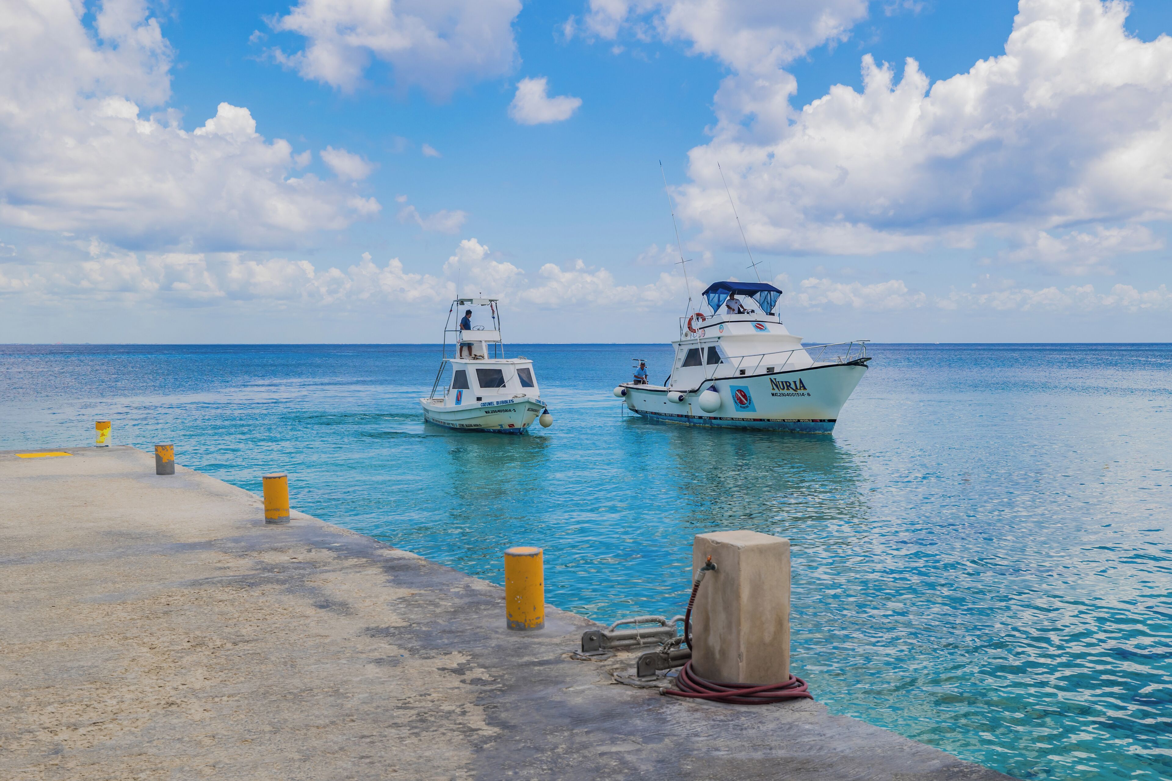 Two boats docked at a pier with clear blue water and a partly cloudy sky.