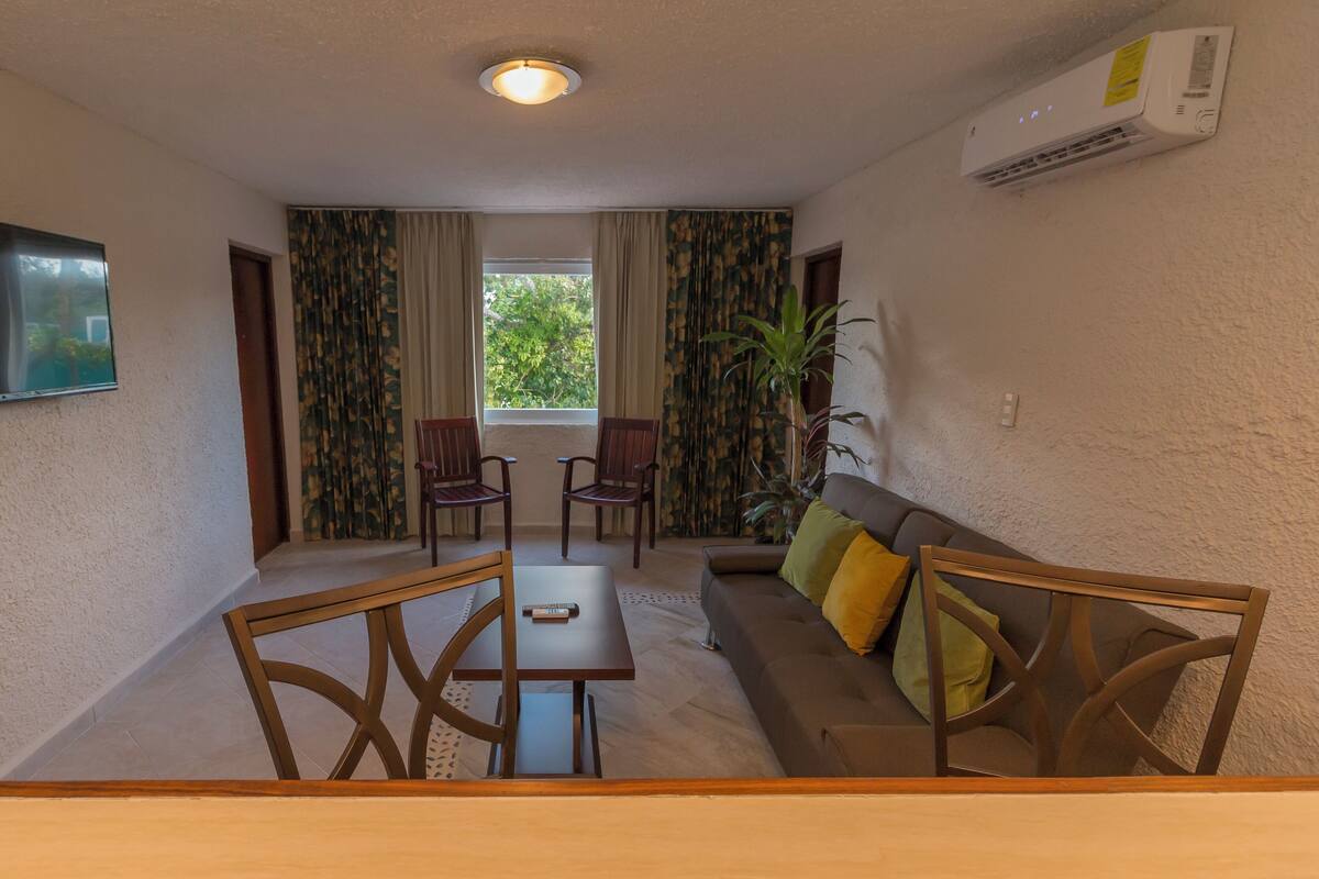 Un salon comprenant un canapé, deux fauteuils, une table, un téléviseur et une fenêtre avec des rideaux.