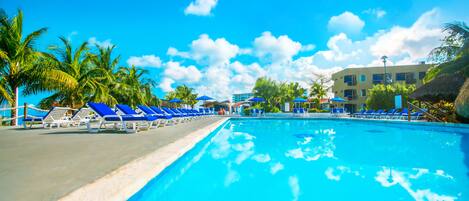 A swimming pool with blue lounge chairs and umbrellas, surrounded by palm trees and a building with multiple windows.