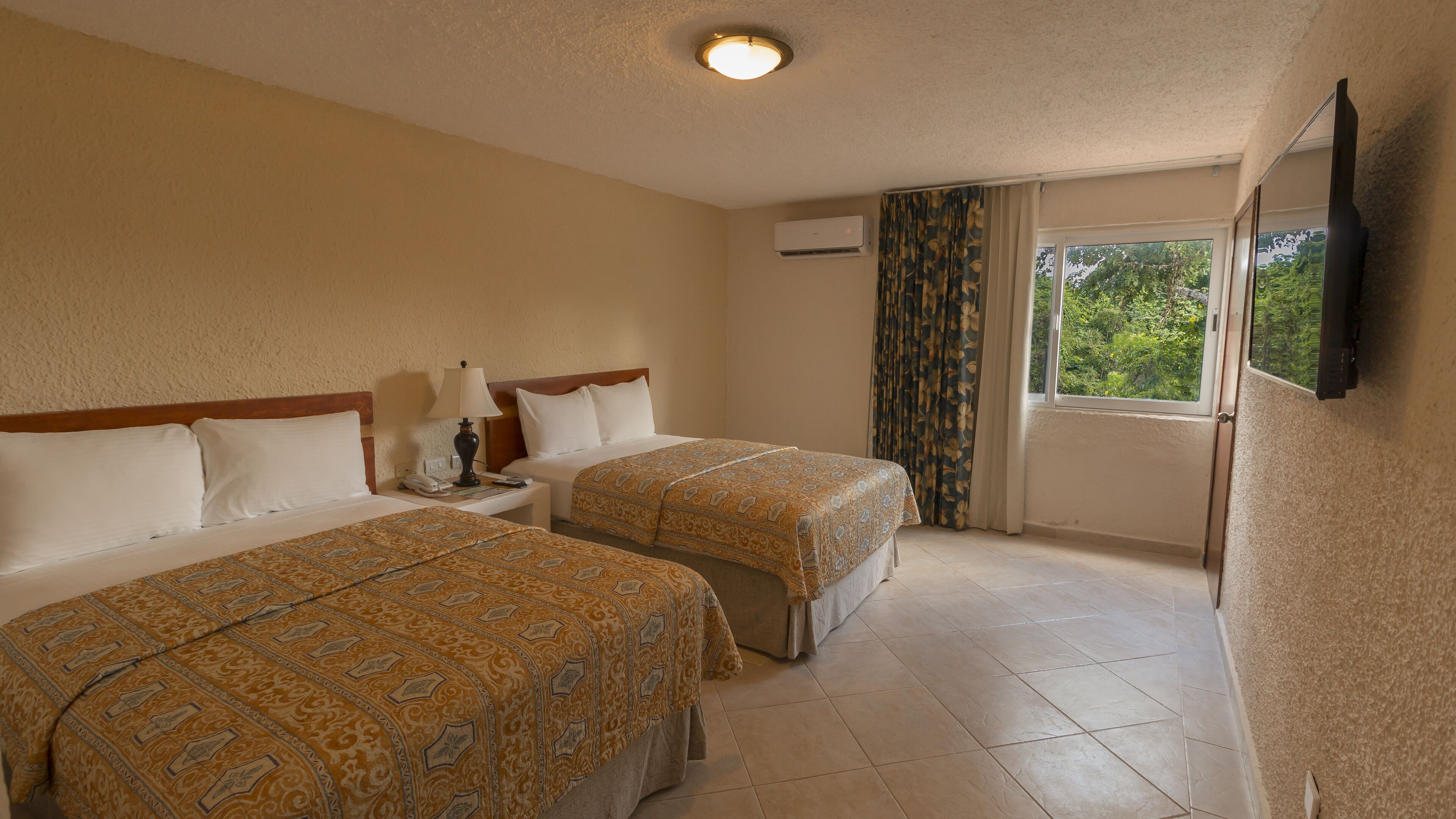 A hotel room with two beds, a nightstand, a lamp, an air conditioning unit, a window with curtains, and a flat-screen TV mounted on the wall.