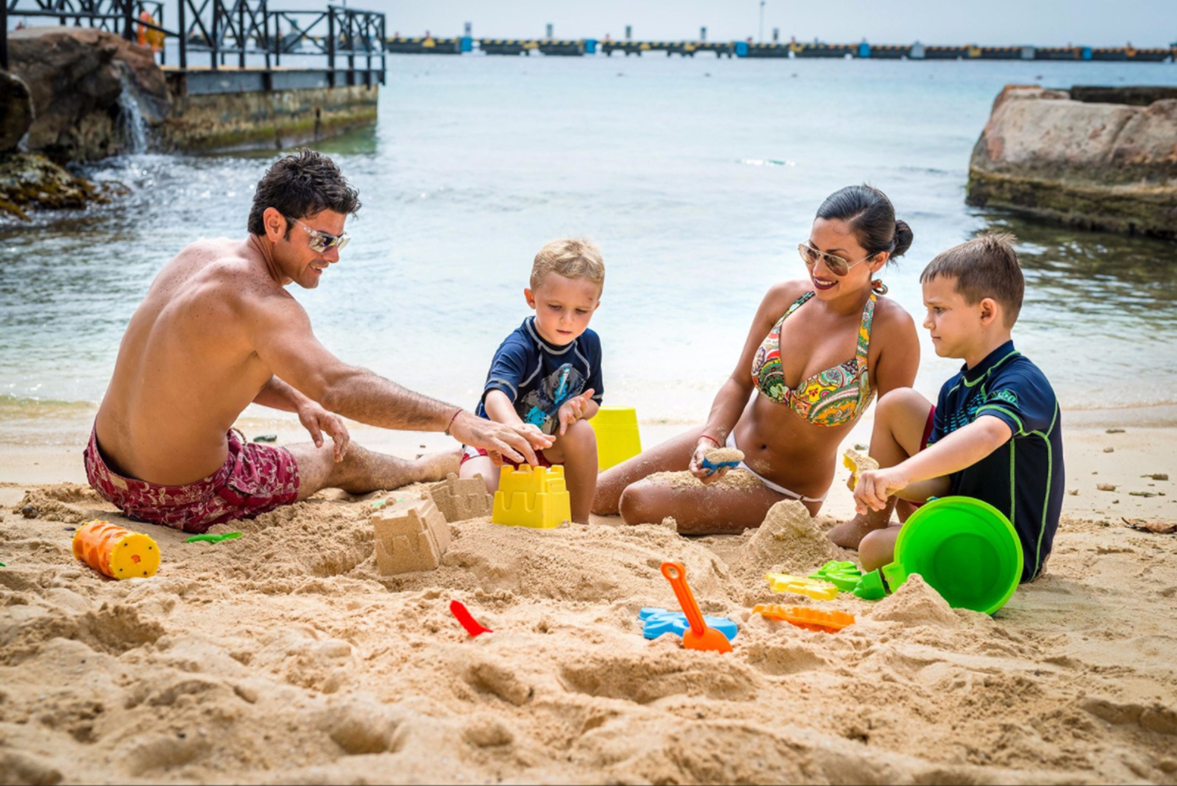 A family of four playing with sand on a beach with a rocky shoreline and a pier in the background.