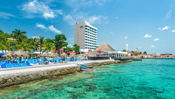 Un hotel frente a la playa con agua turquesa cristalina, un área de piscina con sillas azules y un edificio alto al fondo.