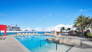 Un área de piscina con sillones de descanso y sombrillas, un gran crucero al fondo y un cielo azul despejado.