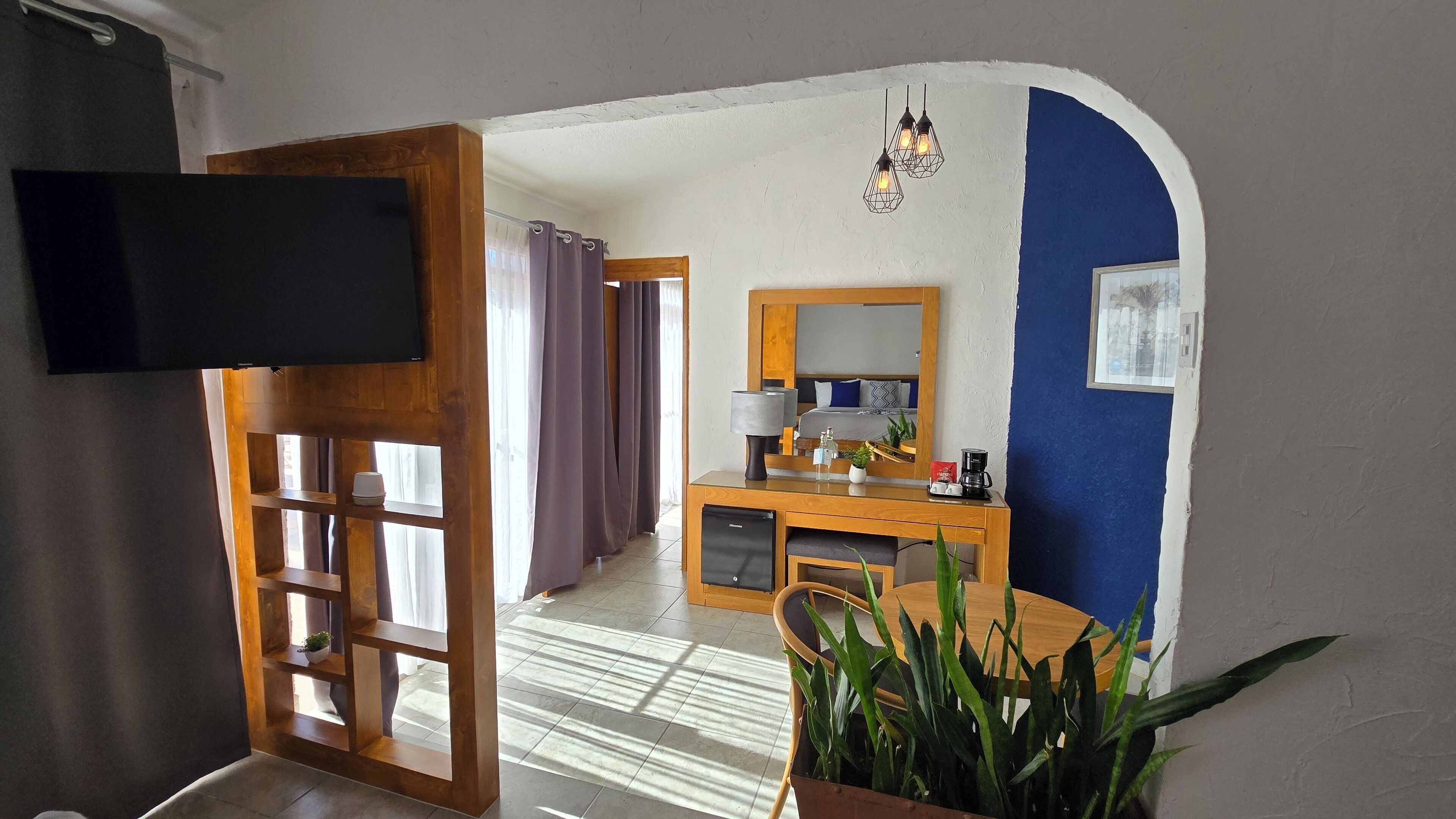 A hotel room with a wooden desk, a mirror, a bed, a TV, and a potted plant.