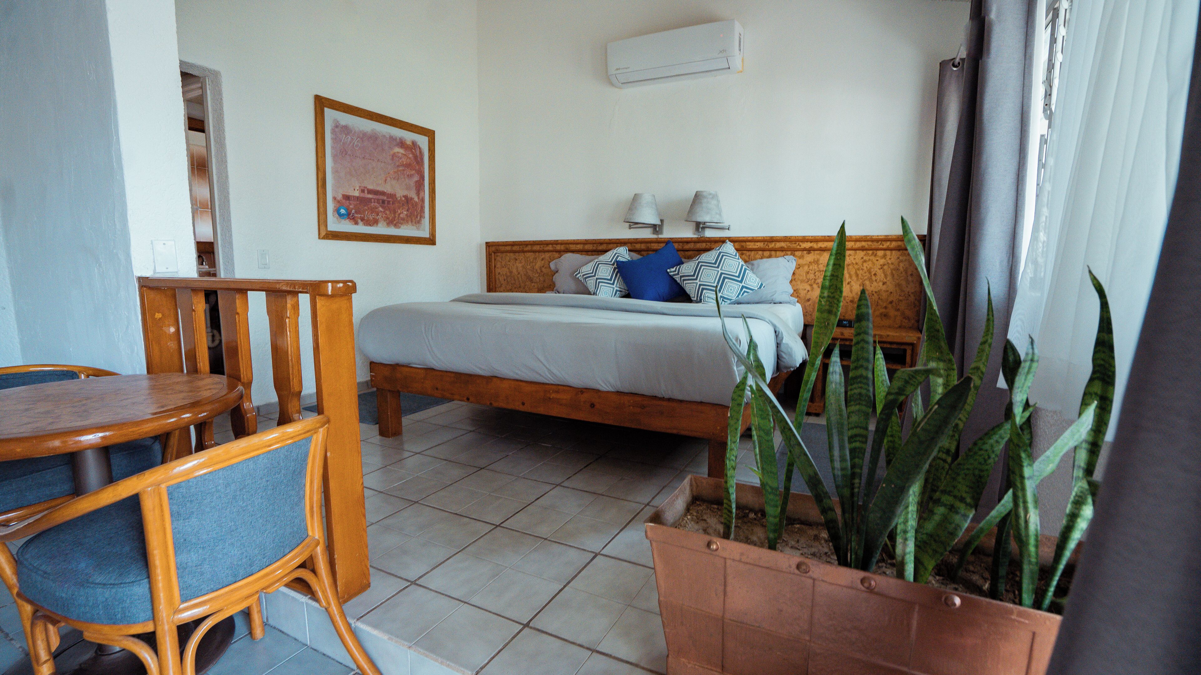 Een kamer met een bed, een houten stoel, een tafel, een kamerplant en een wandairconditioner.