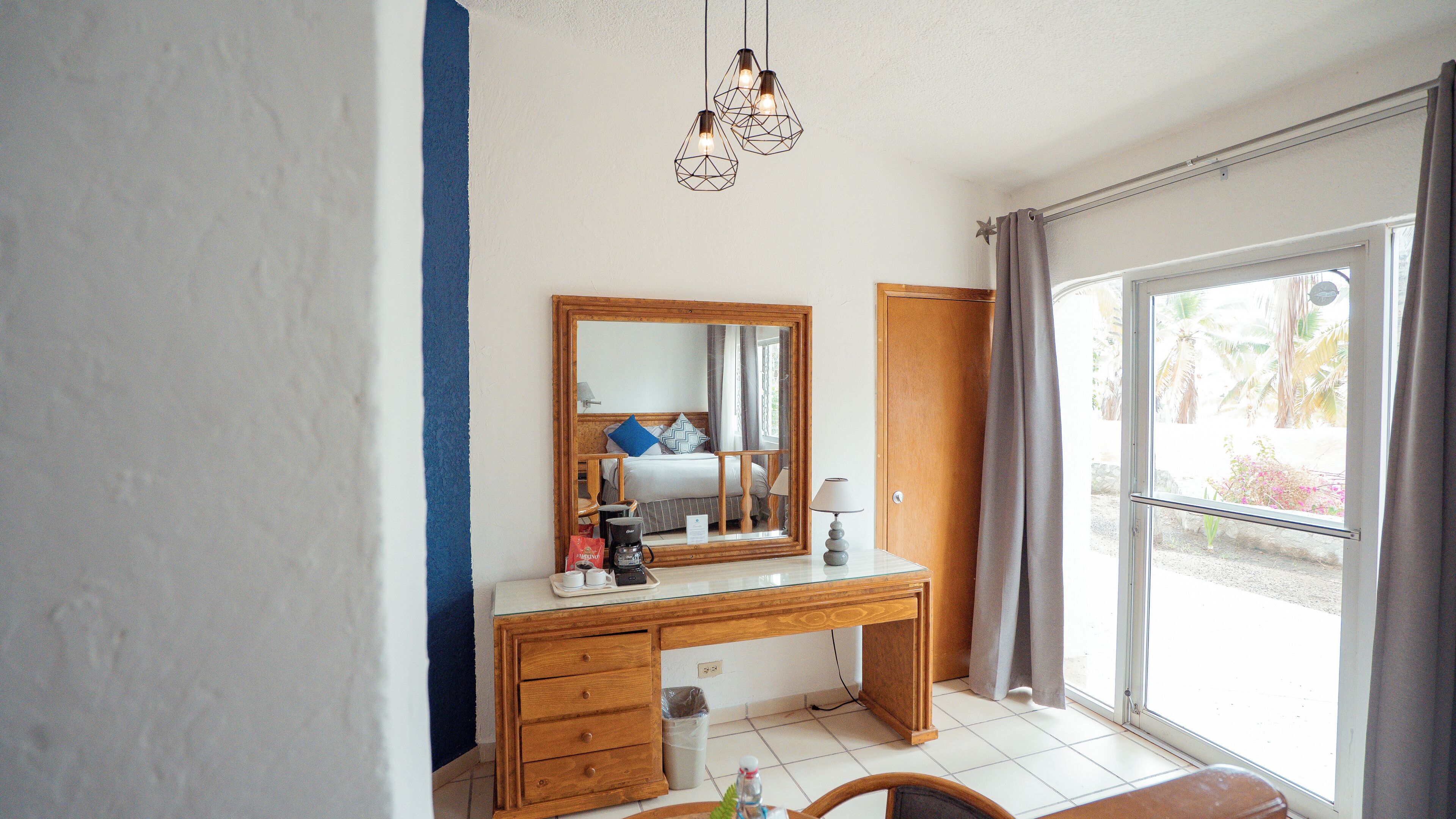A hotel room with a wooden desk, a large mirror, a bed with a view of palm trees, and a balcony with curtains.
