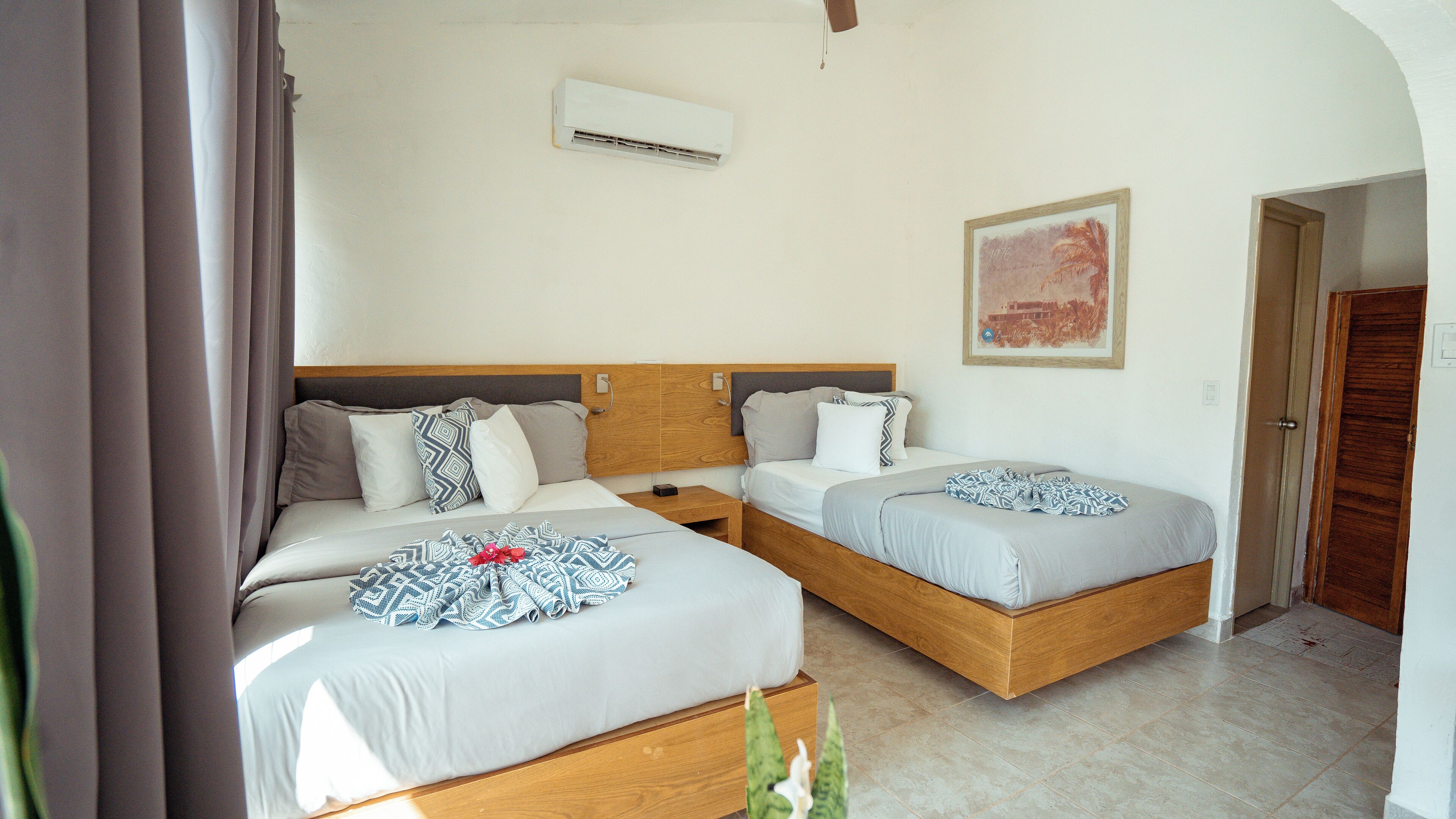 Habitación de hotel con dos camas, cabecero de madera, mesita de noche pequeña, equipo de aire acondicionado y un cuadro en la pared.