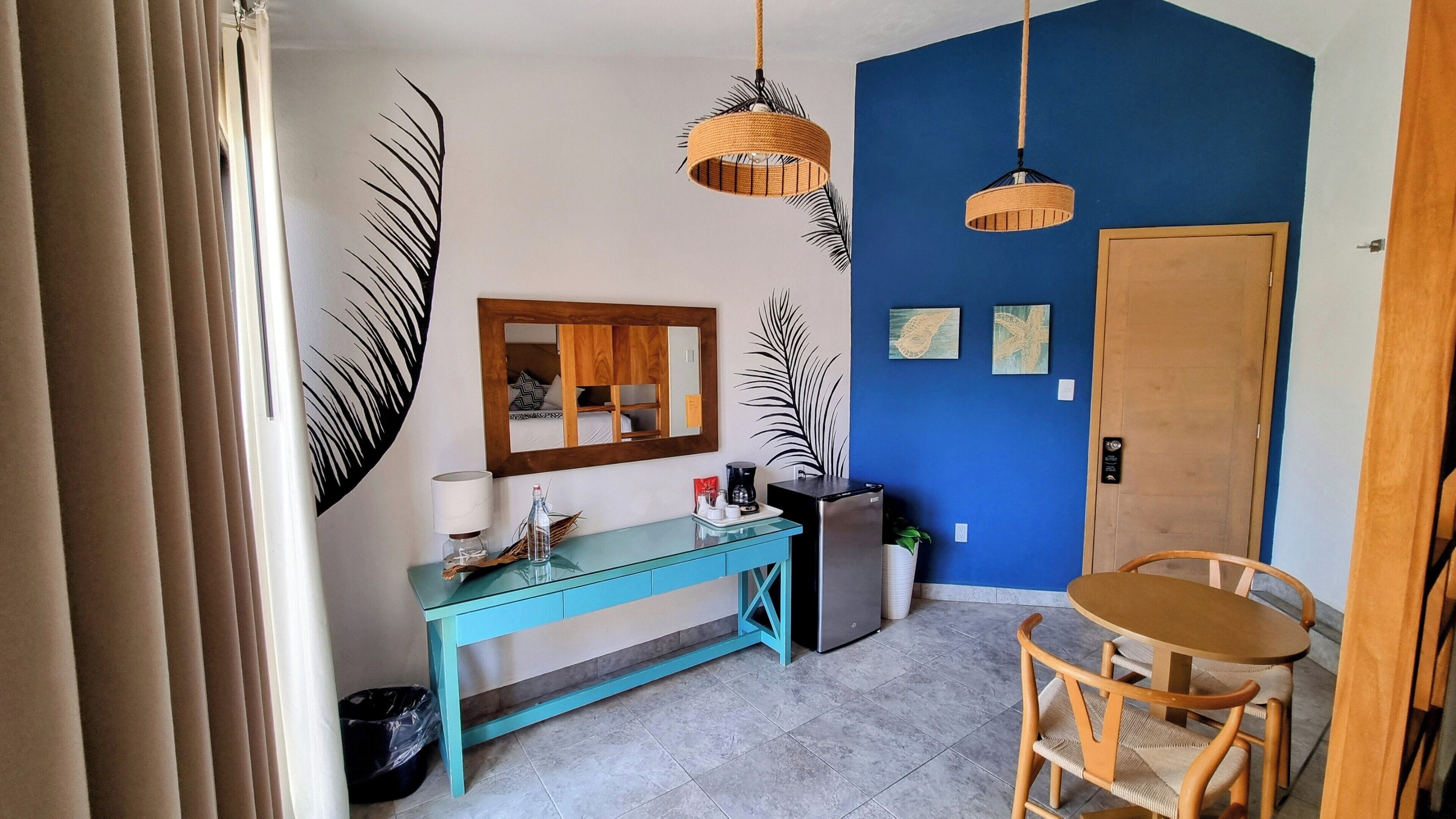 Une pièce avec un mur d’accent bleu, une porte en bois, une table ronde avec des chaises, une console turquoise agrémentée d’un miroir et deux lampes suspendues.