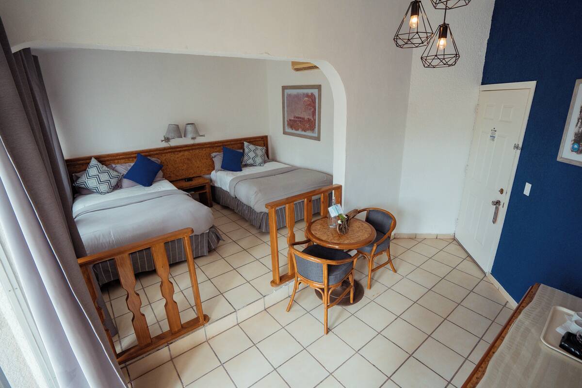 A small, well-lit room with two beds, a wooden table with chairs, and a door.