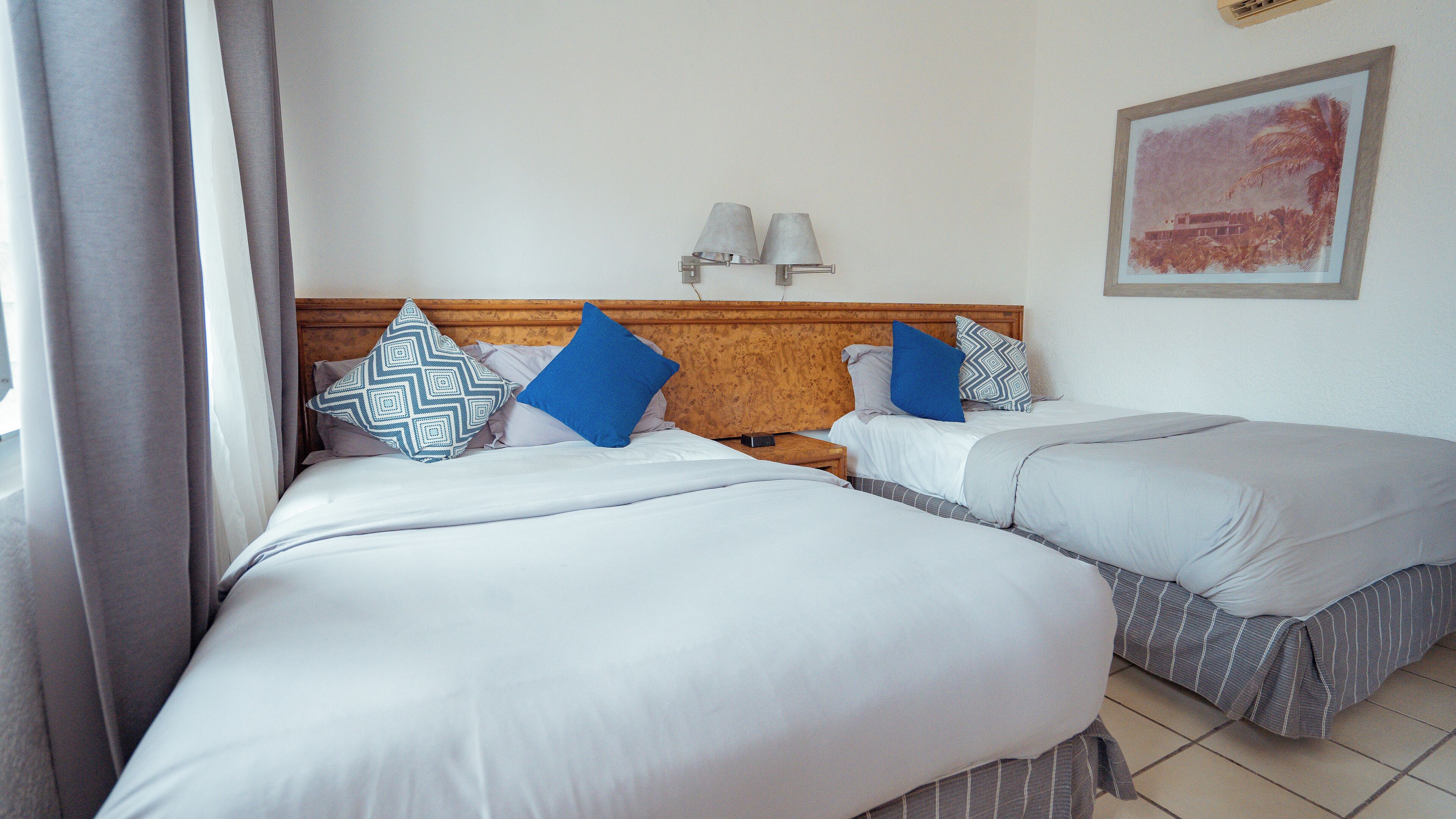 A hotel room with two beds, each with a wooden headboard, white bedding, and patterned pillows. There are two wall-mounted lamps and a framed picture on the wall.