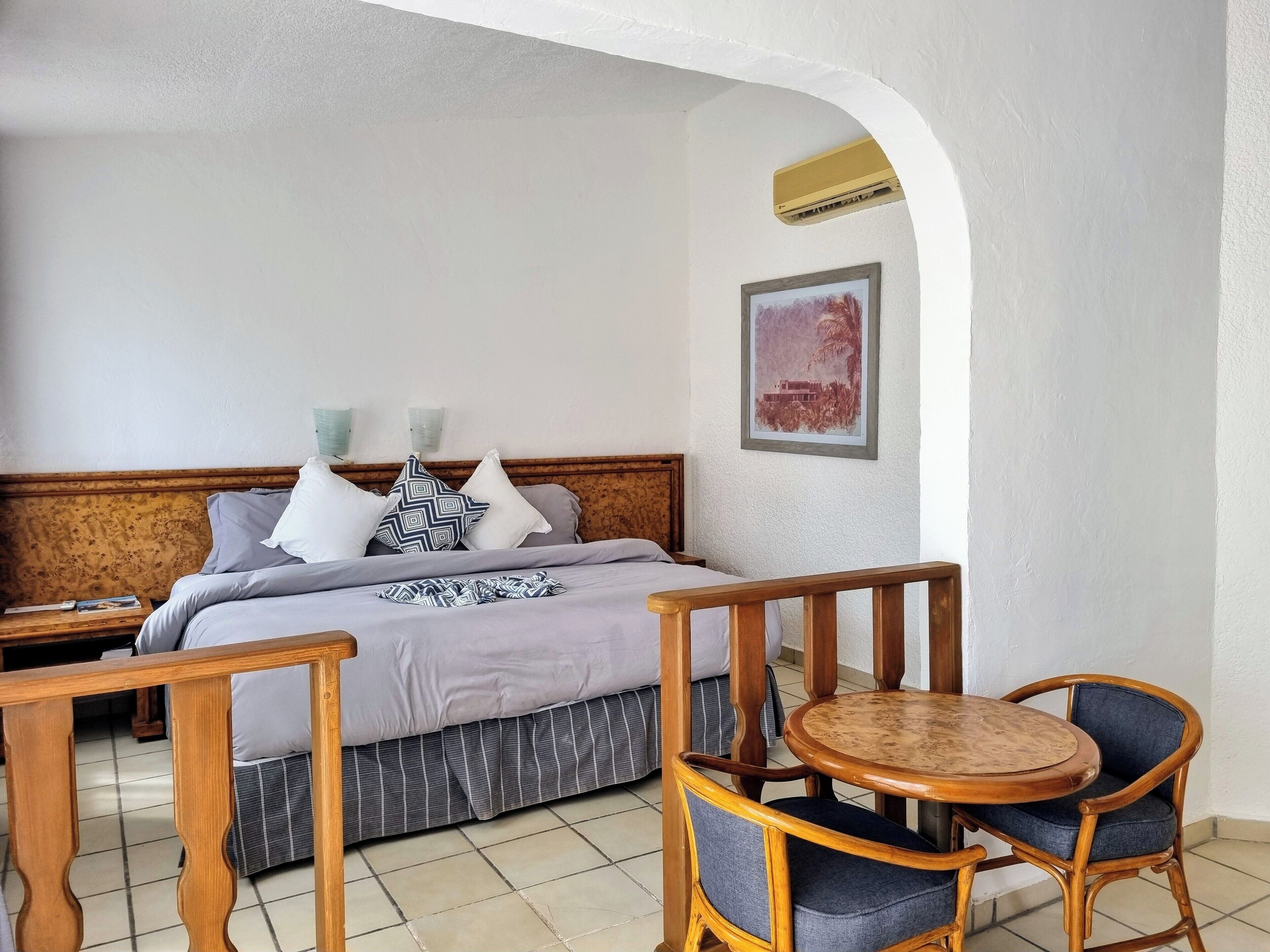 Un dormitorio con cama, cabecera de madera, mesita de noche, pequeña mesa redonda con dos sillas y suelo de baldosas.
