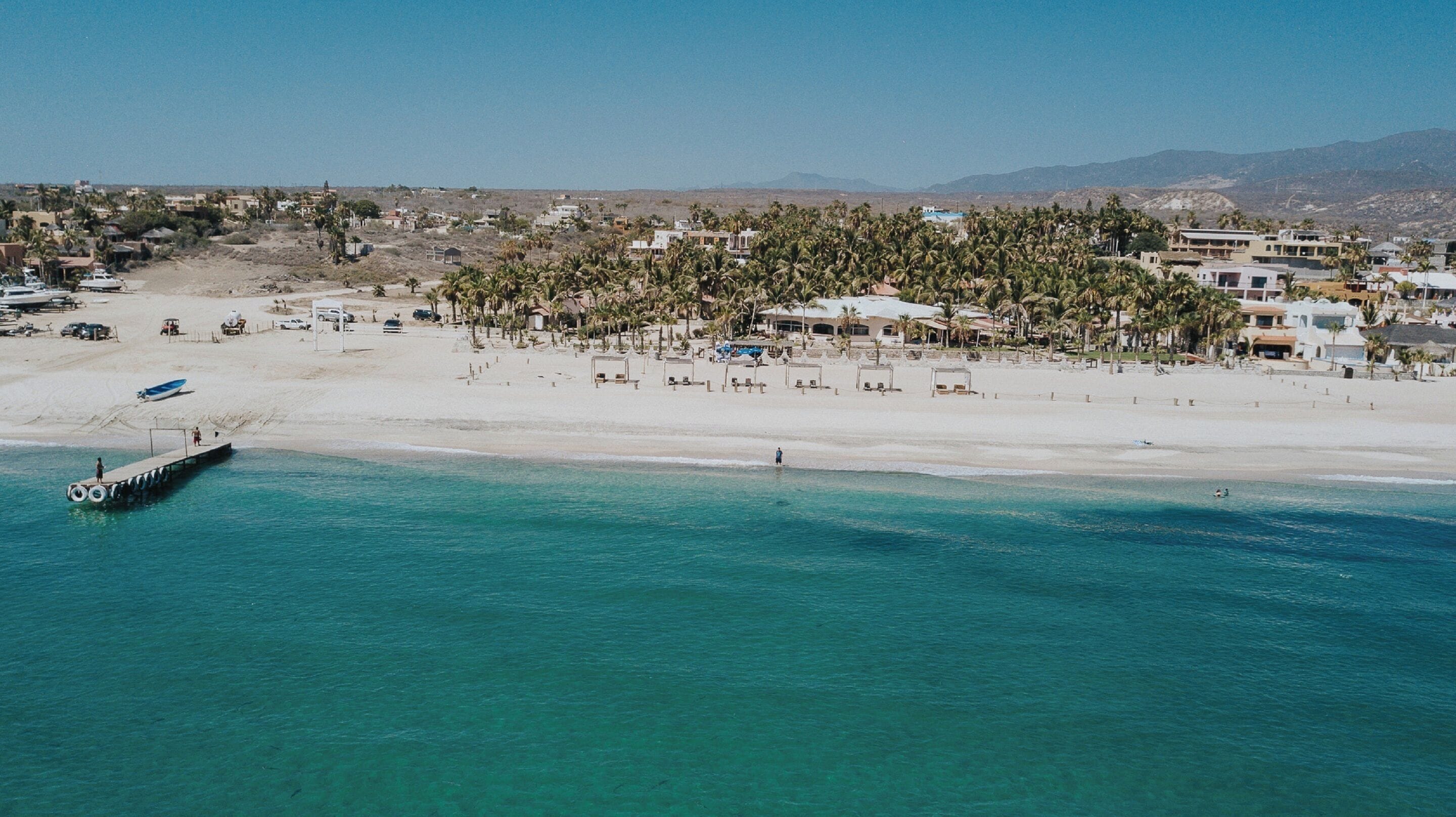 An aerial view of a coastal area with a beach, palm trees, and buildings.