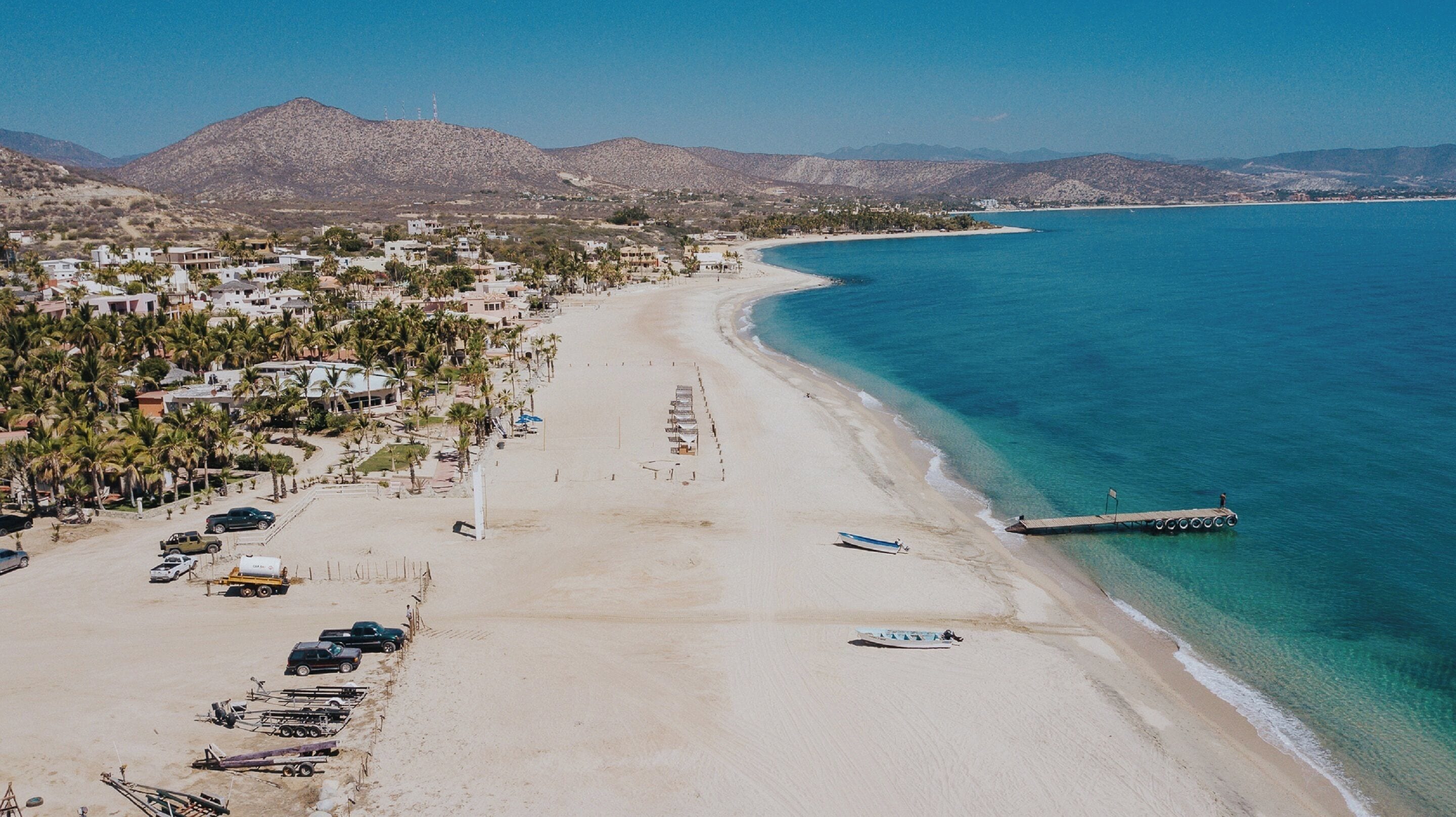 Vista aérea de una playa con una extensa franja de arena, un muelle que se adentra en el agua y un pueblo con edificios y palmeras al fondo.
