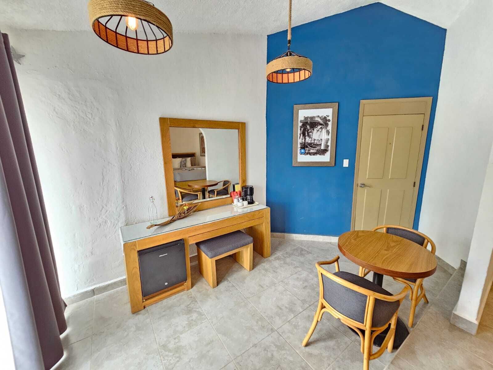 A room with a wooden desk, a mirror, a small refrigerator, a dining table with chairs, and a blue accent wall.