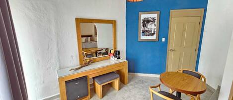 A room with a wooden desk, a mirror, a small refrigerator, a dining table with chairs, and a blue accent wall.