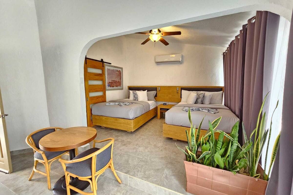 A compact room with two beds, a ceiling fan, an air conditioner, a dining table with chairs, and a potted plant.