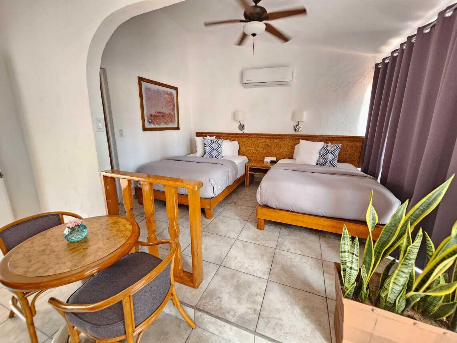 A hotel room with two single beds, a wooden table with chairs, a ceiling fan, and a potted plant.
