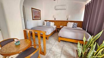 A hotel room with two single beds, a wooden table with chairs, a ceiling fan, and a potted plant.