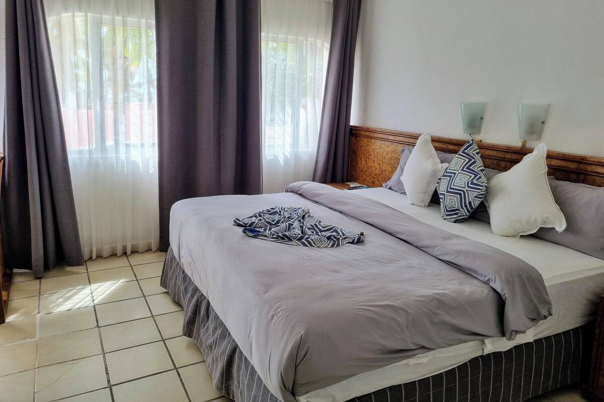 A neatly made bed with a patterned blanket, a headboard with two wall-mounted lights, and a view through sheer curtains to the outside.