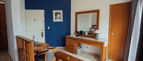 Una habitación con un escritorio de madera, una silla, una pequeña mesa de comedor, un espejo y una puerta.