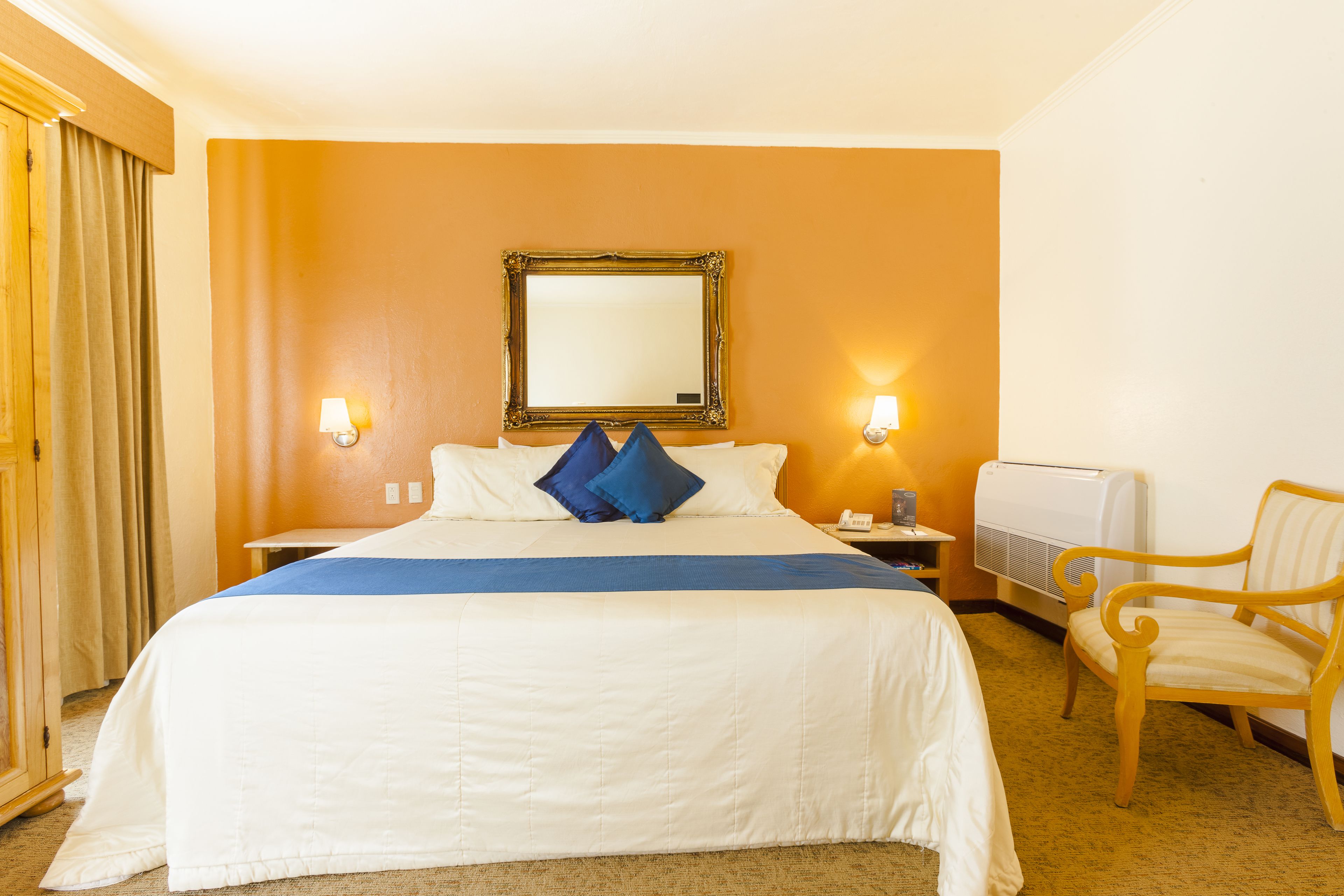 A hotel room with a large bed, two bedside lamps, a chair, and an air conditioning unit.