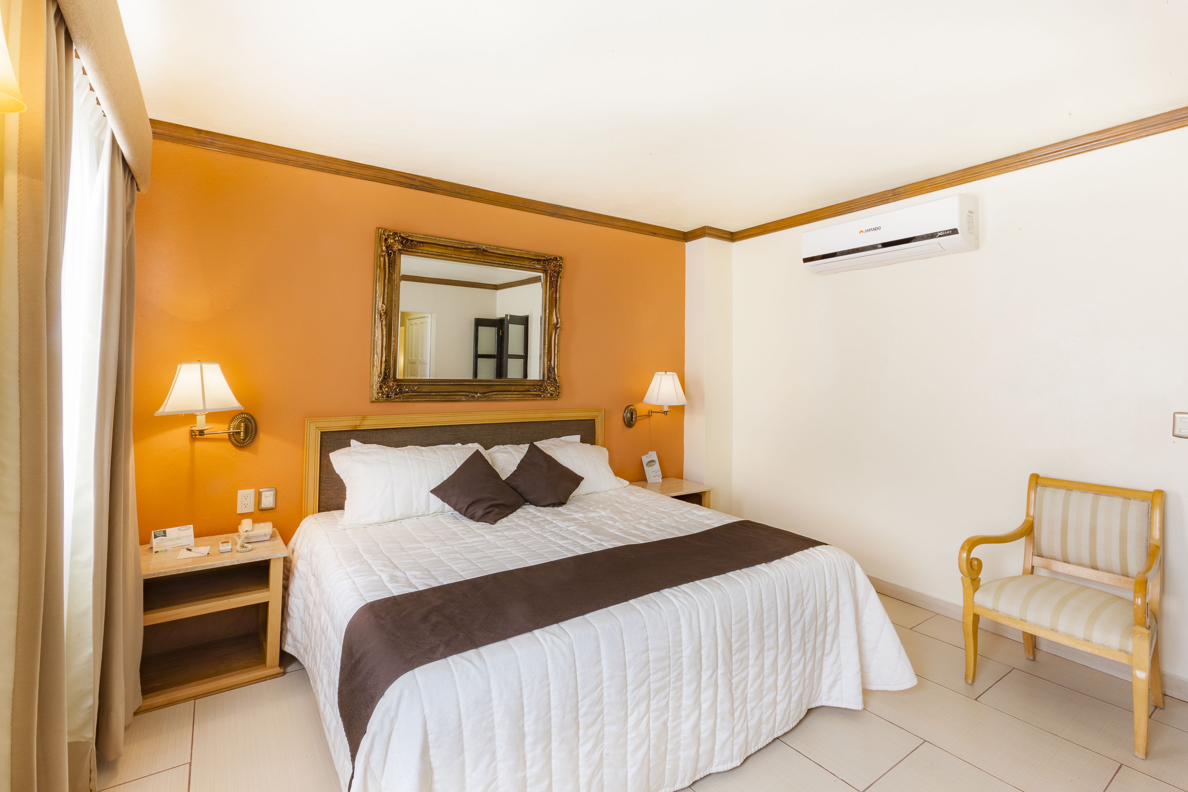Una camera d'hotel con un letto grande, due comodini con lampade, una sedia in legno, uno specchio e un condizionatore d'aria.
