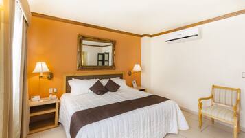 Una camera d'hotel con un letto grande, due comodini con lampade, una sedia in legno, uno specchio e un condizionatore d'aria.