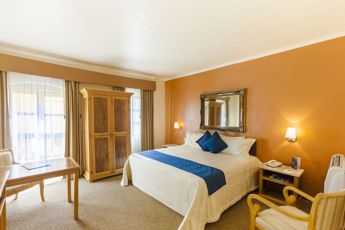 Une chambre d’hôtel comprenant un lit, une table de chevet, une chaise, un bureau et une grande fenêtre avec des rideaux.